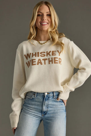 Woman wearing a cream sweater with 'WHISKEY WEATHER' text and blue jeans against a gray background